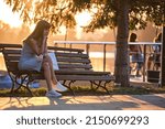 Lonely sad woman sitting alone on lake shore bench on warm summer evening. Solitude and relaxing in nature concept.