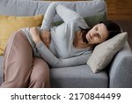 Small photo of Sick young woman suffering from menstrual pain. Woman with hands squeezing belly having painful stomach ache or period cramps sitting on sofa, Abdominal pain, gastritis, and painful periods concept