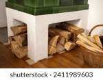 Small photo of Green tiled stove with white base and wood supply - partial view. Next to it there is a basket with kindling wood. Wooden floor