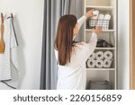 Happy young woman is neatly placing rolled up white towels into a metal mesh basket near bath sink in bathroom. Bath towels storage solution and space saving concept. Personal hygiene and care.