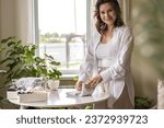 Small photo of Smiling young plus size woman folding linen nightdress in PVC container and metal basket storage organizing. Beautiful female sorting organic cotton textile on table comfortable housekeeping