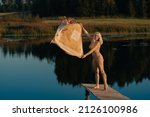 Small photo of A sympathetic blonde girl in a yellow bathing suit poses on a bridge by the lake with a blanket in her hands. Have fun outdoors by the lake next to the forest on a warm summer day.