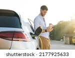 Gorgeous bearded caucasian man in a blue shirt standing near an electric car that is charging and making time adjustments on a smartphone.