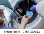 Woman pours liquid washing gel into plastic cap against background of drum of steel-colored washing machine. A girl carefully pours a transparent conditioner for flattening laundry