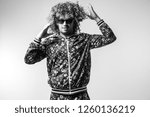 Small photo of Excited cheerful bizarre man posing om white studio background. Stylish funky male with curly hair black and white portrait. Funny guy in tracksuit expressing feelings. Unusual person in sunglasses.