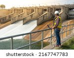 A dam engineering doing his checking routine. He is wearing a white hard hat and yellow transparent vest. He is standing by the rail by the dam.