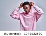 Close up of young african american man in pink streetwear hoodie with hood on head posing isolated on grey background in studio. People lifestyle concept. Copy space. Looking aside down