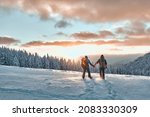 Romantic couple of tourists dressed in warm winter sportswear with tourist backpacks walking with trekking poles in the snowy pine mountains in an incredible sunset. Family, rest.
