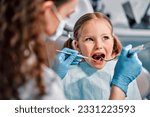 Small photo of Children's dentistry. First examination at the dentist. A cute beautiful girl with an open mouth is looking to the side while the doctor is treating her teeth.