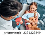 Small photo of A little girl sits in a dental chair, holds a toy rabbit in her hands, laughs and looks at the dentist who shows her a model of the jaw with teeth. Children's dentistry.