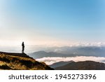 man standing at the edge of the cliff looking at mountains. clouds below