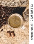 Small photo of From above, a clear cup brimming with coffee sits amidst scattered coffee beans and burlap sacks, creating a captivating tableau of rich textures.