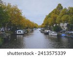 Small photo of Amsterdam, Netherlands; 10, 23, 2022: Trees of feeble leaves along a canal with floating houses and boats