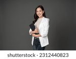 Small photo of Asian female executive with long hair standing hugging a tablet for work, holding a pen, with a beautiful smile. wearing a white suit and stand to take pictures with a gray scene in the studio