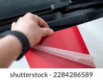 Small photo of A man's hand gluing self-adhesive stripes on a car from red foil. Fast stripe decals on car.
