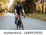 Professional cyclist in sport clothing and protective helmet actively riding bike on paved road among green trees. Strong man in mirrored glasses workout on nature.