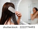Attractive woman in white towel with comb brushing her wet hair after showering at home in front of bathroom mirror. Cares about healthy and clean hair. Beauty concept