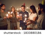 Small photo of Group of cheerful young people in cone party hats with lit sparklers in hands wishing happy birthday to their friend whos about to blow candles on his birthday cake during celebration at home