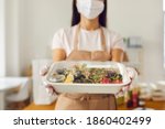 Takeaway cafe worker wearing medical face covering mask holding fresh healthy lunch in plastic food container ready for safe delivery. Concept of ordering meal, good service and taking care of clients