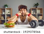 Small photo of Upset depressed young male athlete looking at dumbbell and tempting burger choosing either sport or fast food. Weight loss, fitness, will power, choice between healthy and unhealthy lifestyle concept