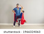 Small photo of Happy strong young dad and active little child dressed as superheroes having fun at home. Father and son in red super hero capes playing together in studio. Family relationship, power concept