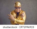 Small photo of Retired older adult man in thug life glasses, gold chain and disco outfit isolated on gray background. Studio portrait senior pensioner in funny sunglasses looking at camera with angry face expression