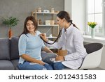 Female doctor listens to the heart and breath of a young woman through a stethoscope on her chest. Woman sitting on sofa at home. Concept of medical care and calling the doctor home.