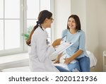 Doctor discussing treatment with cheerful smiley female patient. Happy physician and young woman talking and laughing sitting on examination bed in modern clinic or hospital
