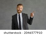 Small photo of Cheerful friendly businessman standing on a gray background hints to someone that he is cool. Stylish bearded man in a suit winks and points to the side. Concept of success and business relationships.