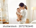 Mother holding baby. Happy beautiful young mom standing in bedroom or nursery room interior in tender white and pastel colors, holding cute sweet little child and smiling. Family, love, care concept