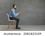 Small photo of Side view portrait of a serious businessman sitting tense at an imaginary desk with a computer. Man keeps his hands outstretched as if typing on a keyboard and working on a laptop. Gray background.