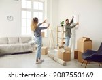 Happy married couple who recently bought house are unpacking stuff and decorating new home. Young man and woman are making living room cozy and choosing place on wall to hang painting or family photo