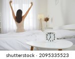 Good morning. Wake up. Rise and shine. It's time to start new working day. White alarm clock that tells 7 AM standing on bedside table, and woman who woke up from night sleep stretching in background