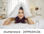 Annoyed and angry woman closes her ears with pillow in morning because of noise. Close up portrait of sleepless woman annoyed by alarm clock or loud neighbors. Concept of insomnia, stress and tinnitus