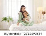 Happy family enjoying lazy morning at home. Portrait of smiling mother and child in bedroom. Beautiful mum and daughter in comfortable white and pastel green lounge homewear cuddling on cosy, warm bed