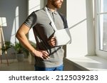 Orthopedic emergency. Injured man who during rehabilitation at home wearing medical support brace and sling bandage immobilizer on his arm. Young man with broken arm looks thoughtfully out window.