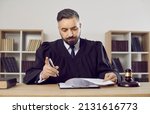 Small photo of Judge working at his desk in court. Portrait of serious judge, lawyer or attorney dressed in robe gown uniform sitting at table with gavel hammer mallet, holding pen and reading judiciary law document