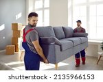 Small photo of Two professional relocation service workers in overalls move sofa in customer's apartment. Movers carry sofa, cardboard boxes and assembling furniture. Moving and delivery company services.