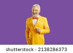 Small photo of Studio shot of happy white haired bearded senior man wearing bright yellow suit, white shirt, orange bow tie and trendy glasses standing with his hand in pocket isolated on solid purple background