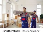 Small photo of Smiling young male movers carrying carpet unload goods in client home or office. Happy removal company male workers unpack help client with boxes during moving or relocation. Transportation.