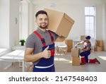 Small photo of Smiling male worker of moving and delivery company holding cardboard box showing thumbs up. Loader in overalls posing against background of colleague who packs cardboard boxes. Moving service concept.