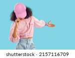 Small photo of Child shows something on blue blank copyspace studio background. Pretty kid invites you to see new collection and go shopping. Funky Black girl in pink bubble gum ball cap presents fashion novelty