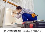 Small photo of Plumber fixing problems in the kitchen. Man in an overall work uniform crouching on the floor beside the sink drain, trying to unclog the blockage, and checking, repairing or replacing some pipe parts