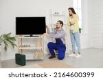 Young friendly service man installs and connects new TV in female client's house. Young woman watches as male repairman in work uniform connects cables to TV screen. Service and maintenance concept.
