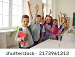 Small photo of Happy clever school children ready for class. Small group of cheerful cute smart kids in casual clothes with books and backpacks standing near row of desks in classroom, raising up hands and smiling