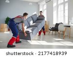 Small photo of Two young workers lift up heavy sofa together. Young men from moving company and lorry delivery service removing things from the house, carrying furniture and other belongings