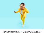 Full body happy cheerful Afro American woman wearing stylish yellow suit and trendy glasses standing isolated on blue background, looking at camera, winking her eye and smiling. Party, fashion