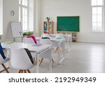 Small photo of Bright interior of empty classroom without students in modern elementary school. School classroom with blackboard, white desks and chairs with colored backpacks. Educational concept.