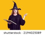 Small photo of Serious creepy woman in witch character pointing with finger to side on orange background in studio. Portrait of woman with bloody makeup in black dress and witch's hat holding spell book. Web