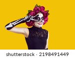 Happy beautiful woman with skull face makeup and flowers on head does peace victory sign with two fingers, smiles and looks at camera isolated on yellow background. Halloween, Day of the Dead concept